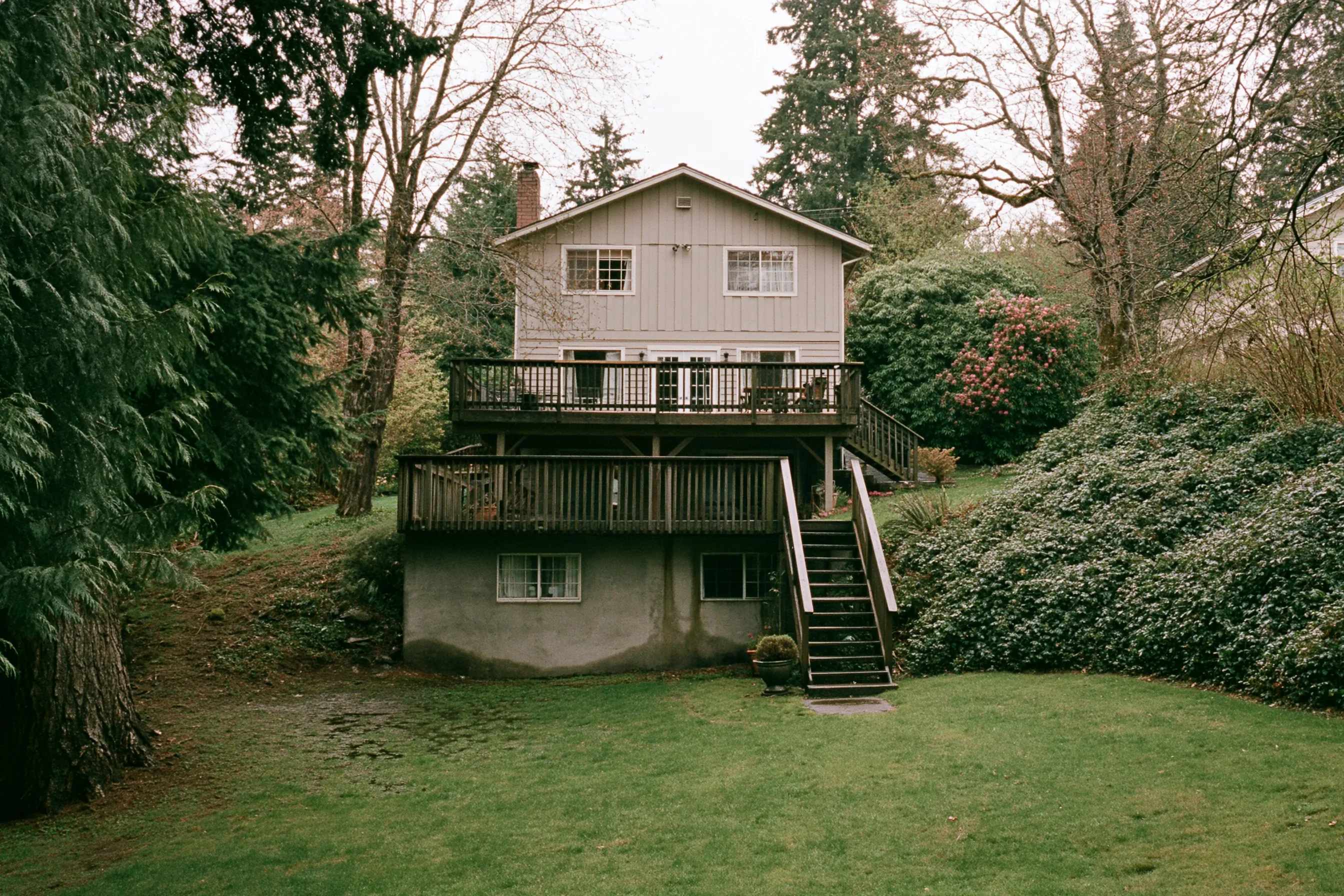 Rear of a two-story house on a gentle slope with a daylight basement, a poured concrete wall visible at the lower level, a deck above, wet lawn and Douglas firs